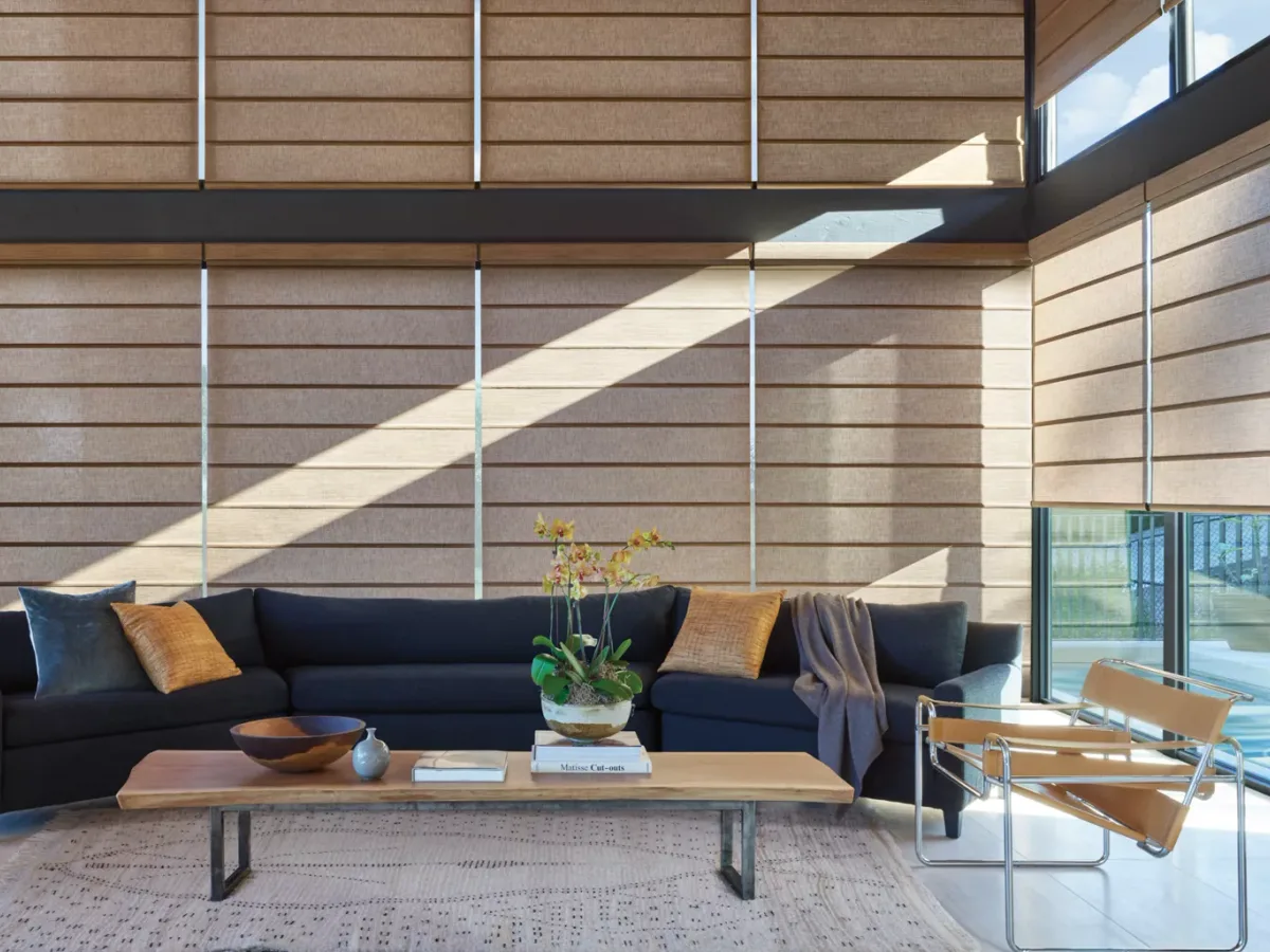 A two-storey wall of Hunter Douglas Alustra Woven Textures Roman shades in an architectural living room, sunlight raking across the sculptural fabric folds