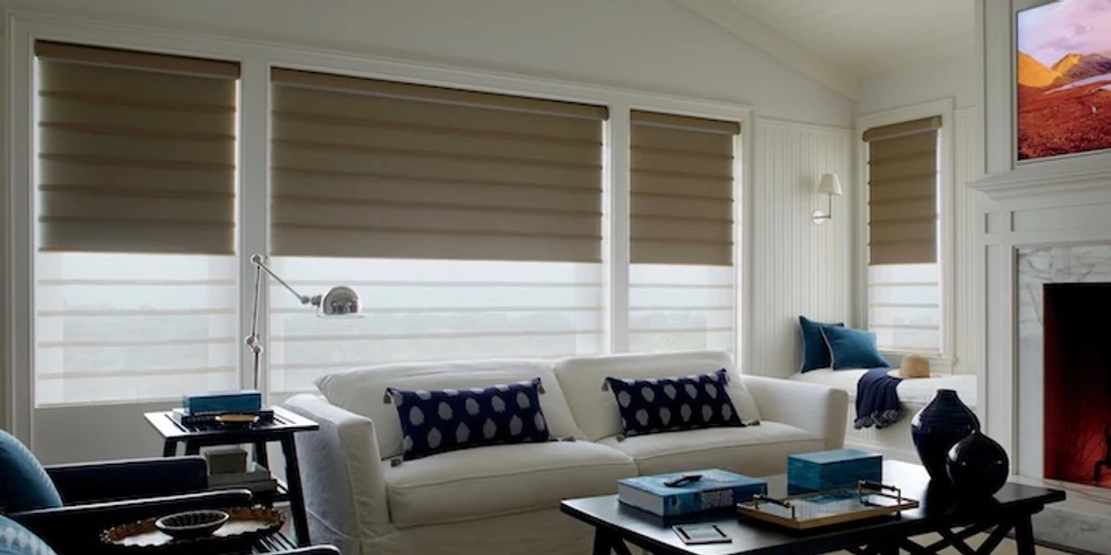 Hunter Douglas Vignette Duolite shades across a living room with the opaque back shade lowered halfway: the dark room-darkening fabric covers the top of each window and the pale light-filtering sheer glows across the bottom