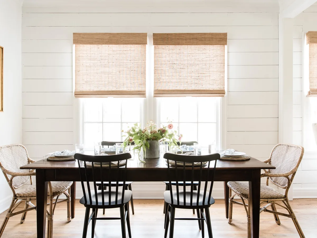 Hunter Douglas Provenance woven wood Roman shades over two windows in a white shiplap dining room, daylight glowing through the natural weave